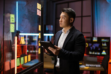 Kanban scrum board, project management, work efficiency concept. Pensive Korean businessman using tablet computer, managing project tasks, working near office glass board with sticky notes