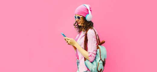 Summer colorful portrait of stylish modern young woman listening to music in headphones with smartphone on pink background