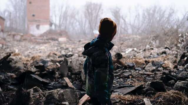 A boy walks through the ruins of destroyed houses under the falling snow. He looks sad and he's looking for someone.