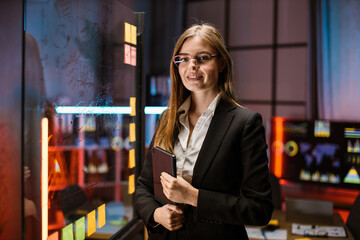 Confident smiling pretty female ceo executive manager wearing glasses and formal clothes, holding tablet computer, standing near glass wall with sticky papers, planning project in modern office