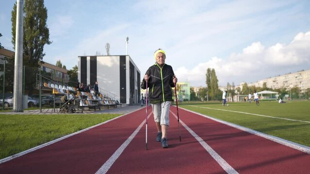 Senior woman walking with walking poles in stadium on a red rubber cover. Elderly woman 88 years old doing Nordic walking exercises at the city stadium on the running track. Healthy lifestyle concept.