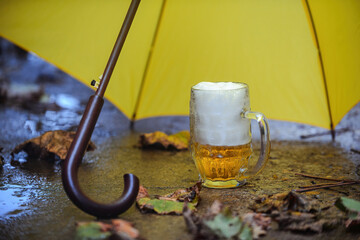 on the street on a rainy autumn cloudy day, a bright yellow umbrella stands on the ground, under the umbrella there is a beer mug with a foamy alcoholic drink beer, fallen autumn leaves are around