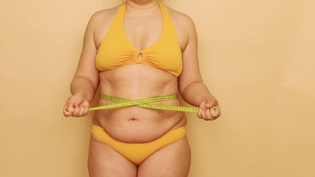 Unrecognizable fat overweight woman standing in yellow bra, bikini, showing excess belly, tightening waist with tape on beige background. Body positive, cellulite, obesity, weight control, measuring.