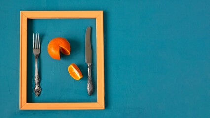 Pop art style food. A modern creative concept of a fruit diet. Tangerine in a pink wooden frame with a knife and fork on a sea-green surface.