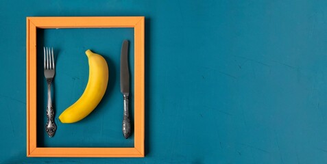Pop art style food. A modern creative concept of a fruit diet. Banana in a pink wooden frame with a knife and fork on a sea-green surface.