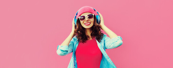 Colorful portrait of happy laughing young woman listening to music in headphones wearing a casual hat on pink background
