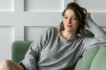 Beautiful young woman relaxed on the couch in the living room at home. Daylight. Female in a grey sweater filling comfortable at her house. Weekend