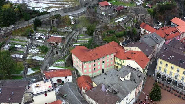 Aerial drone footage of a medieval castle in Europe.  High castle walls and towers, with the village below.  Surrounded by snowy mountains with an overcast sky. 
