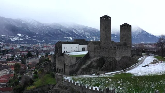 Aerial drone footage of a medieval castle in Europe.  High castle walls and towers, with the village below.  Surrounded by snowy mountains with an overcast sky. 