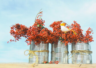 Recycled cans with red berries. Three recycled cans with bunches of red berries (Schinus terebinthifolia) with an artificial bird outdoors and blue sky.