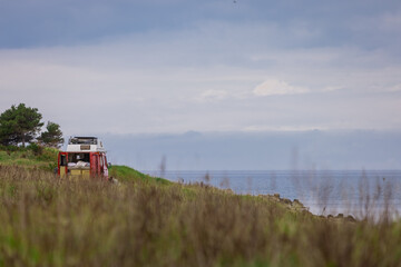Fototapeta premium Vanlife or van life in its fullest. Red vintage old van with visible bed in the back parked in the middle of nature overlooking the sea on a cloudy day. Freedom with a van.