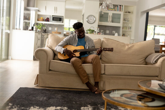 Smiling african american young man playing guitar while sitting on sofa in living room at home