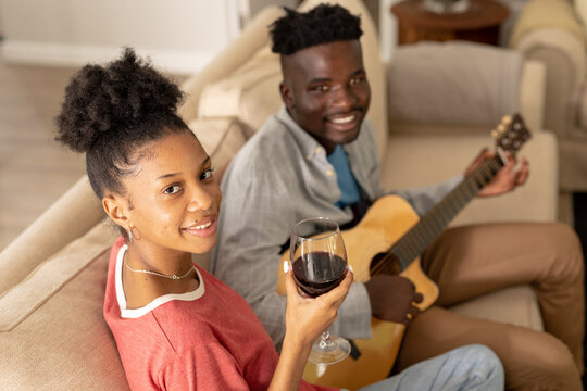 Portrait of smiling african american young woman holding wine and boyfriend playing guitar on sofa