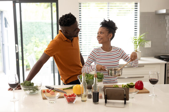 Smiling african american young man looking at girlfriend preparing food in kitchen at home