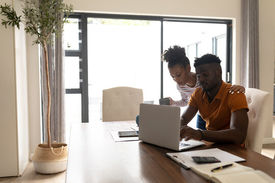 African american young woman standing by boyfriend working over laptop on table at home