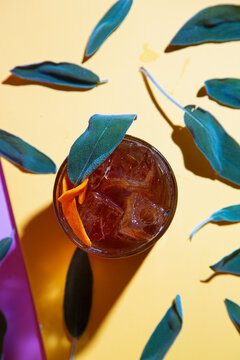 Overhead shot of brown cocktail on the rocks with sage leaves and orange garnish