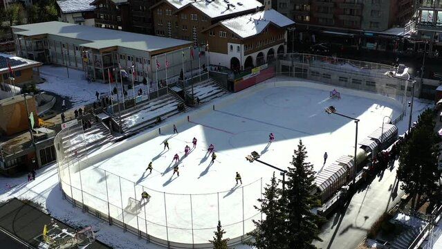 Aerial drone footage from Zermatt, Switzerland.  A game of ice hockey is being played in the middle of the city, on a sunny day.  