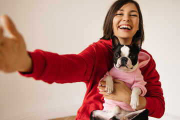 Cute caucasian young woman holding dog on her lap takes photo with french bulldog. Girl with dark hair wears hoodie with her pet. Animal care concept.