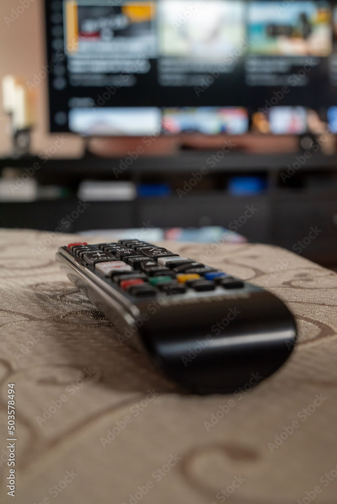 Poster closeup shot of a black remote control on the table with big screen t.v in the blurred background