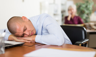 Fototapeta premium Exhausted overworked businessman sleeping on table with laptop and documents, resting his head on hands. Daily work and fatigue concept