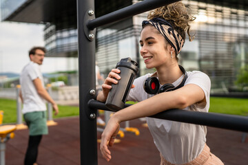 Fototapeta premium One caucasian woman taking a brake during outdoor training in the park outdoor gym resting on the bars with supplement shaker in hand drinking water or supplementation happy smile copy space