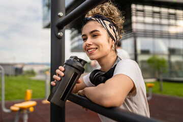 One caucasian woman taking a brake during outdoor training in the park outdoor gym resting on the bars with supplement shaker in hand drinking water or supplementation happy smile copy space