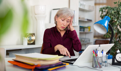 Frustrated tired mature woman working with papers and laptop in office