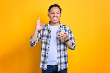 Cheerful young Asian man in plaid shirt using mobile phone and showing okay sign gesture, recommend application isolated on yellow background