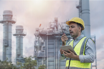 Engineer wearing PPE uniform and helmet looking detail tablet on hand with power plant  on background.