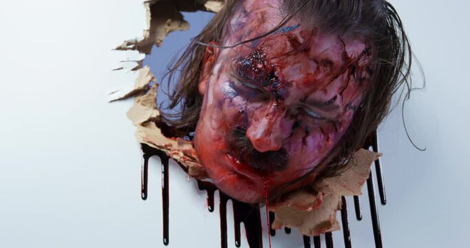 Exhausted zombie put head into hole in white plasterboard wall. Spooky man with red dirty face breaks through with blood flowing out of mouth closeup