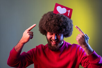 Hipster man holding pointing at Heart Like icon of social media on his head, notification button to follow, subscribe blog. Indoor studio shot isolated on colorful neon light background.