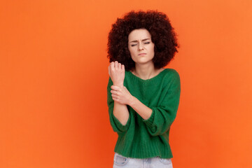 Sad woman with Afro hairstyle wearing green casual style sweater injured wrist, suffering from pain, holding sick hand, keeps eyes closed. Indoor studio shot isolated on orange background.