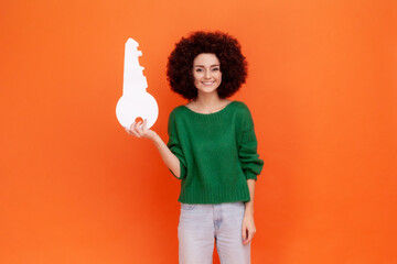 Portrait of smiling woman with Afro hairstyle wearing green casual style sweater holding paper key in hands, looking at camera, property. Indoor studio shot isolated on orange background.
