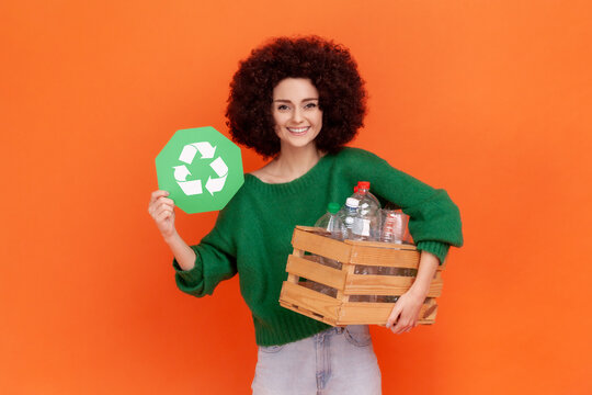 Smiling Woman With Afro Hairstyle Wearing Green Casual Style Sweater Holding Box With Plastic Bottles And Showing Green Recycling Sign. Indoor Studio Shot Isolated On Orange Background.