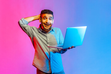 Portrait of man in shirt looking at camera with amazed expression, watching shocking content, surprised by news, raised arm. Indoor studio shot isolated on colorful neon light background.