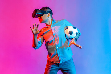 Man in shirt wearing headset, standing with soccer ball and raised arms, watching vr football match, worried for his favorite team. Indoor studio shot isolated on colorful neon light background.
