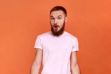 Obraz premium Portrait of amazed bearded man standing with mouth open in surprise, has shocked expression, hears unbelievable news, wearing pink T-shirt. Indoor studio shot isolated on orange background.