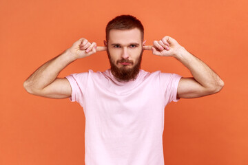 Portrait of bearded man closing ears with fingers seriously looking at camera, tired of irritated noise, wants silence, wearing pink T-shirt. Indoor studio shot isolated on orange background.