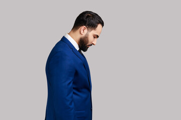Portrait of sad upset bearded man standing with his head down and looking, having with dissatisfied sadness face, wearing official style suit. Indoor studio shot isolated on gray background.