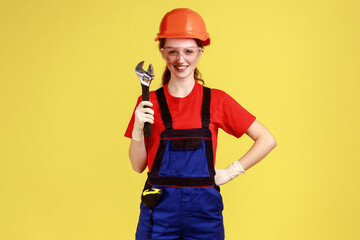 Portrait of smiling satisfied builder woman holding adjustable wrench, looking at camera with happy facial expression, wearing overalls and helmet. Indoor studio shot isolated on yellow background.