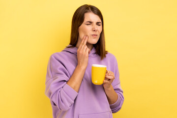 Portrait of sad sick unhealthy woman has teeth pain after drinking hot or cold beverage, dental injury, wearing purple hoodie. Indoor studio shot isolated on yellow background.