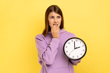 Portrait of impatient nervous attractive woman biting her nails and holding big wall clock, deadline, looking away, wearing purple hoodie. Indoor studio shot isolated on yellow background.