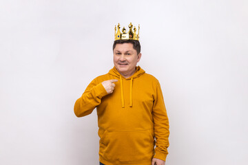 I'm king. Middle aged man in golden crown and pointing himself, looking at camera with smile, superior privileged status, wearing urban style hoodie. Indoor studio shot isolated on white background.