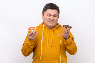 Healthy eating vs junk food. Confused man holding doughnut and fresh grapefruit, looking with...