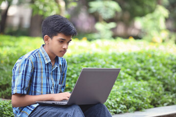 Indian young boy using Laptop while Sitting Outside park	
