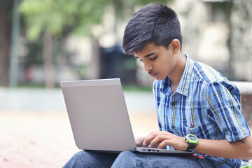 Indian young boy using Laptop while Sitting Outside park	
