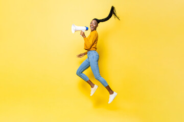Fototapeta na wymiar Portrait of young African American woman holding megaphone jumping in isolated yellow studio background