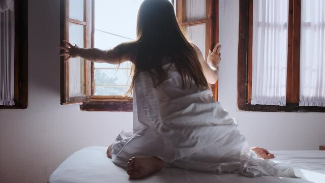 Young woman in white sheet opens window sashes in morning. Long-haired, romantic traveler woke up in old town Berat. Girl in hotel bed. Opening travel destinations of Albania. Travel lifestyle