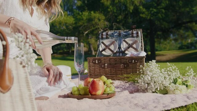 Unrecognizable blonde woman in white boho dress pouring bubbling champagne drink in flute glass for drinking in green park. 6K slow motion RED camera footage with picnic background Video series 5 of 6