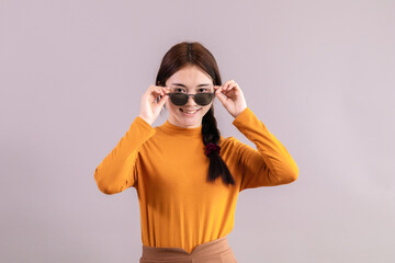 Adorable Asian woman action in front of gray background in studio wearing sun eye glasses. Tourist girl happy acting with camera,  girl in autumn fashion casual yellow and orange pointing finger